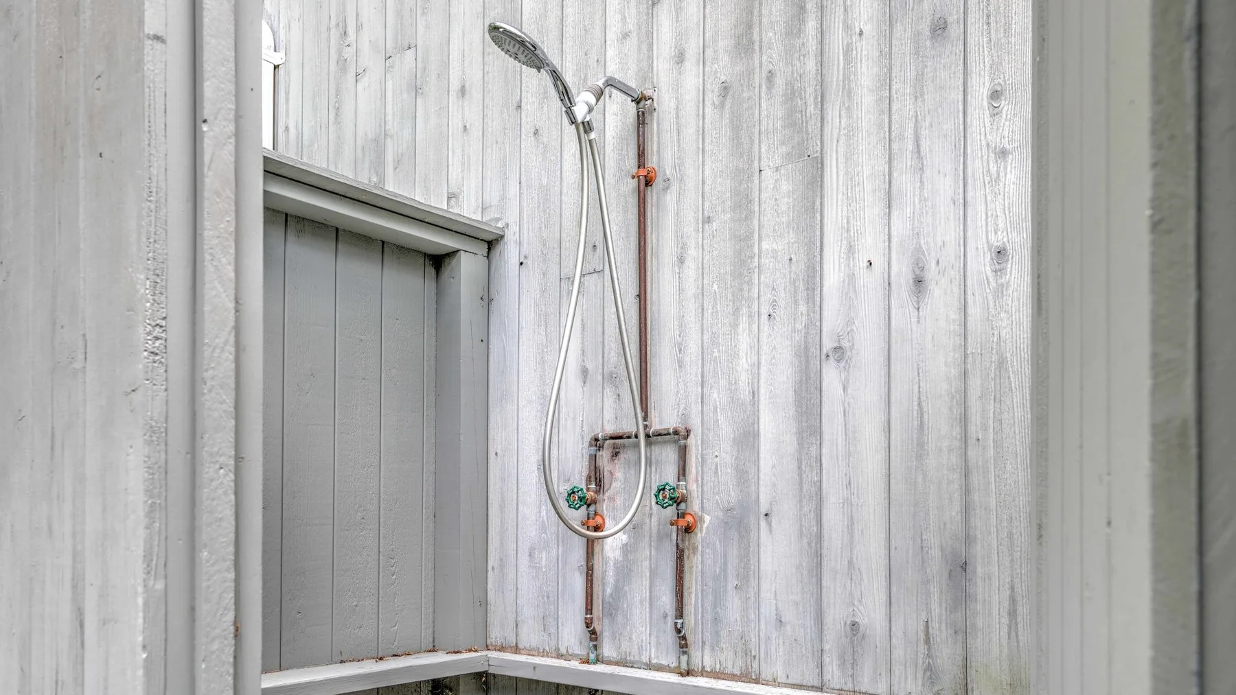 A shower with a handheld showerhead and visible plumbing against pale walls.