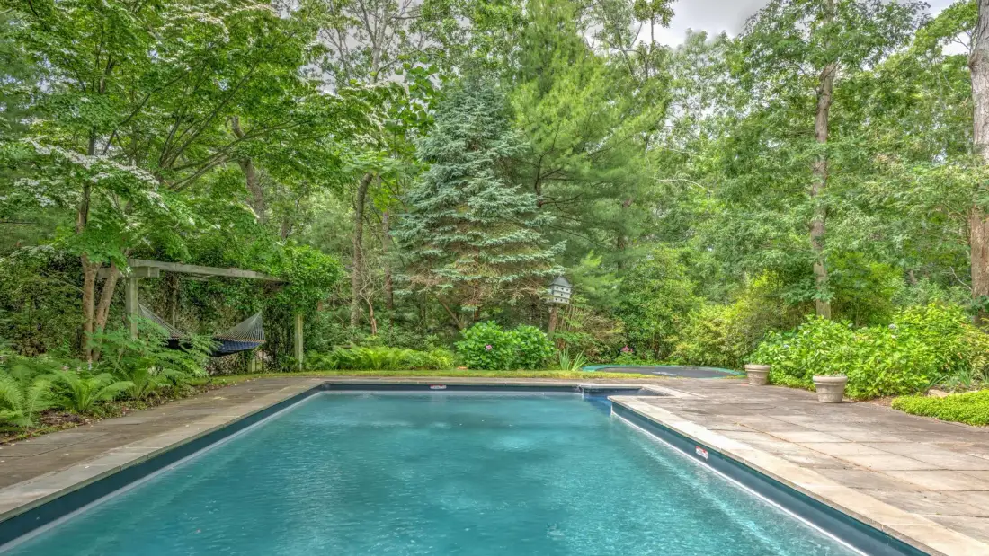 An outdoor swimming pool with a paved deck, surrounded by trees and greenery.