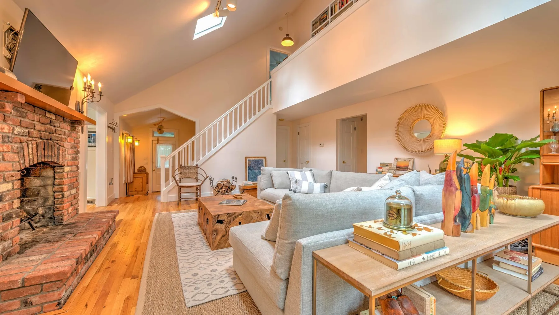 A bright living room with a brick fireplace, pale seating, and a staircase.