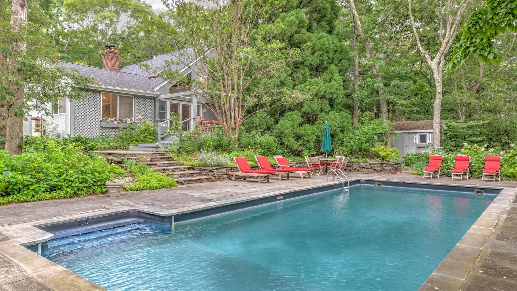 An outdoor swimming pool sits beside a landscaped garden and a house.