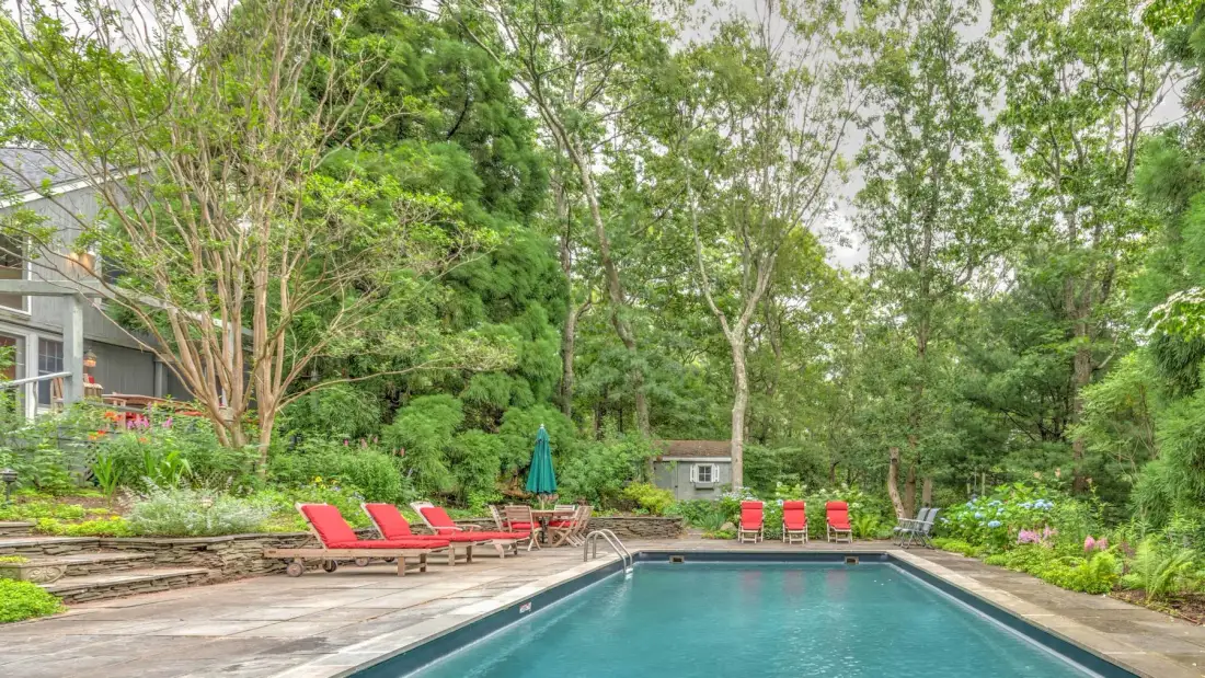 A swimming pool is set in a wooded garden with lounge seating nearby.