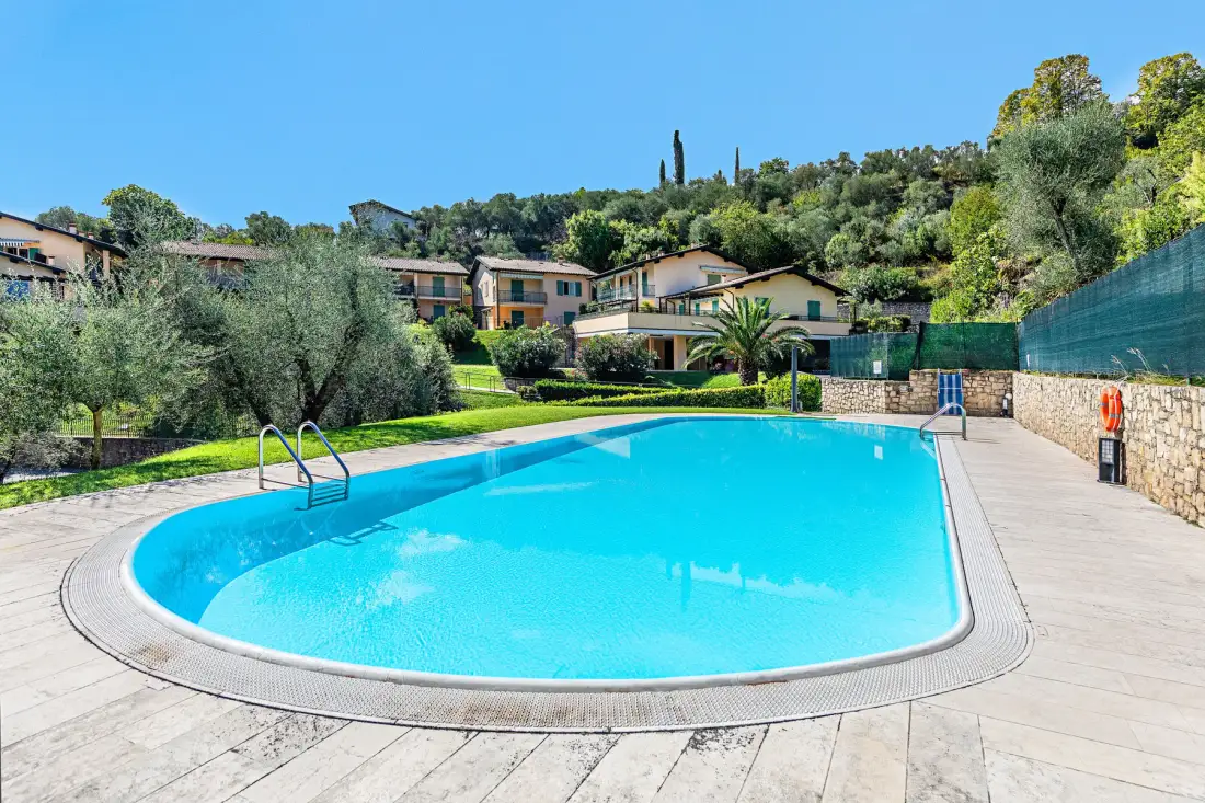 An outdoor swimming pool sits beside a paved deck, with houses and greenery in the background.