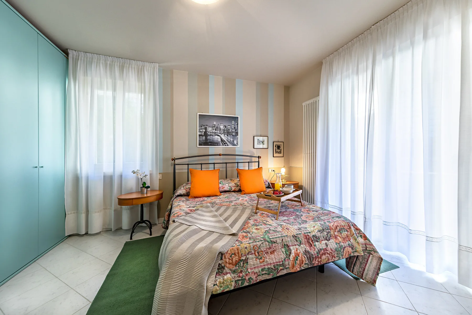 A bedroom with a bed dressed in patterned bedding and tall windows with sheer curtains.