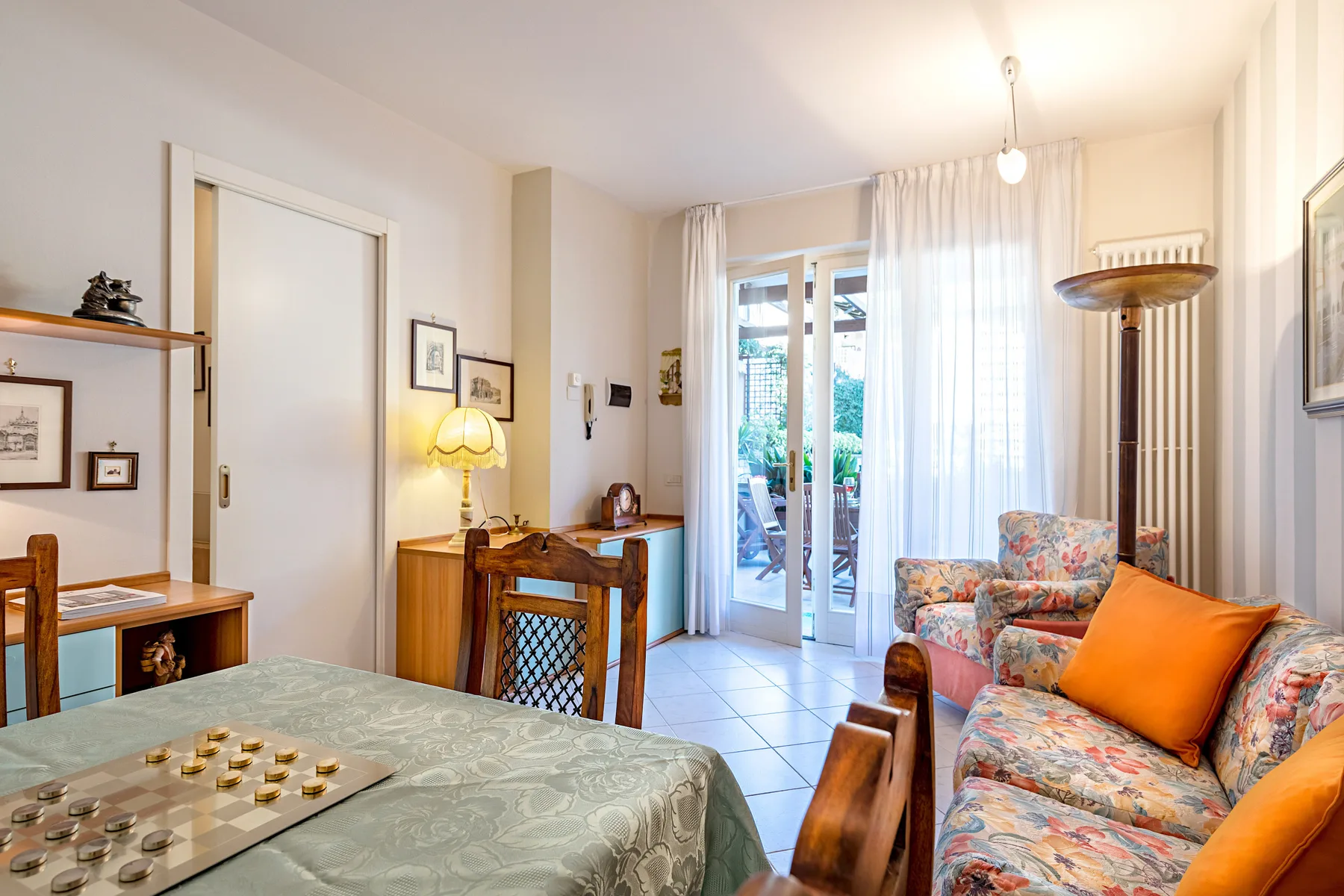 A bright living and dining area has a table, floral seating, and glass doors opening outside.