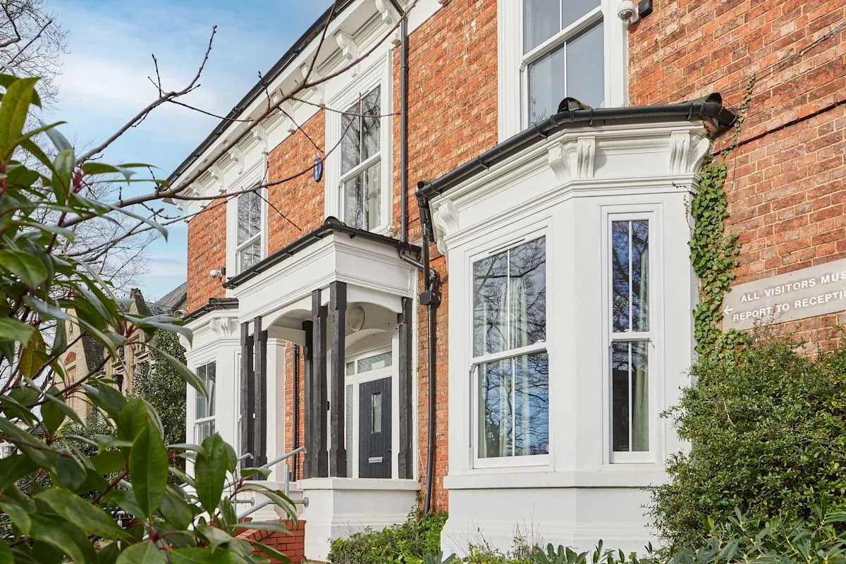 A red-brick building exterior with white-trimmed windows and leafy plants.