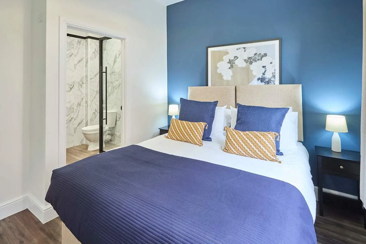 A bedroom with a bed dressed in blue and a glass shower visible through the doorway.
