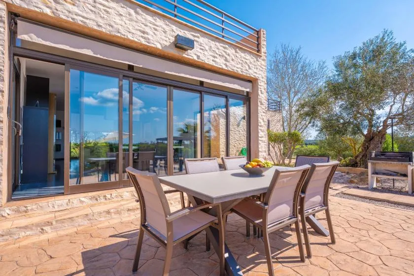 An outdoor dining area sits beside a stone house and large glass doors.
