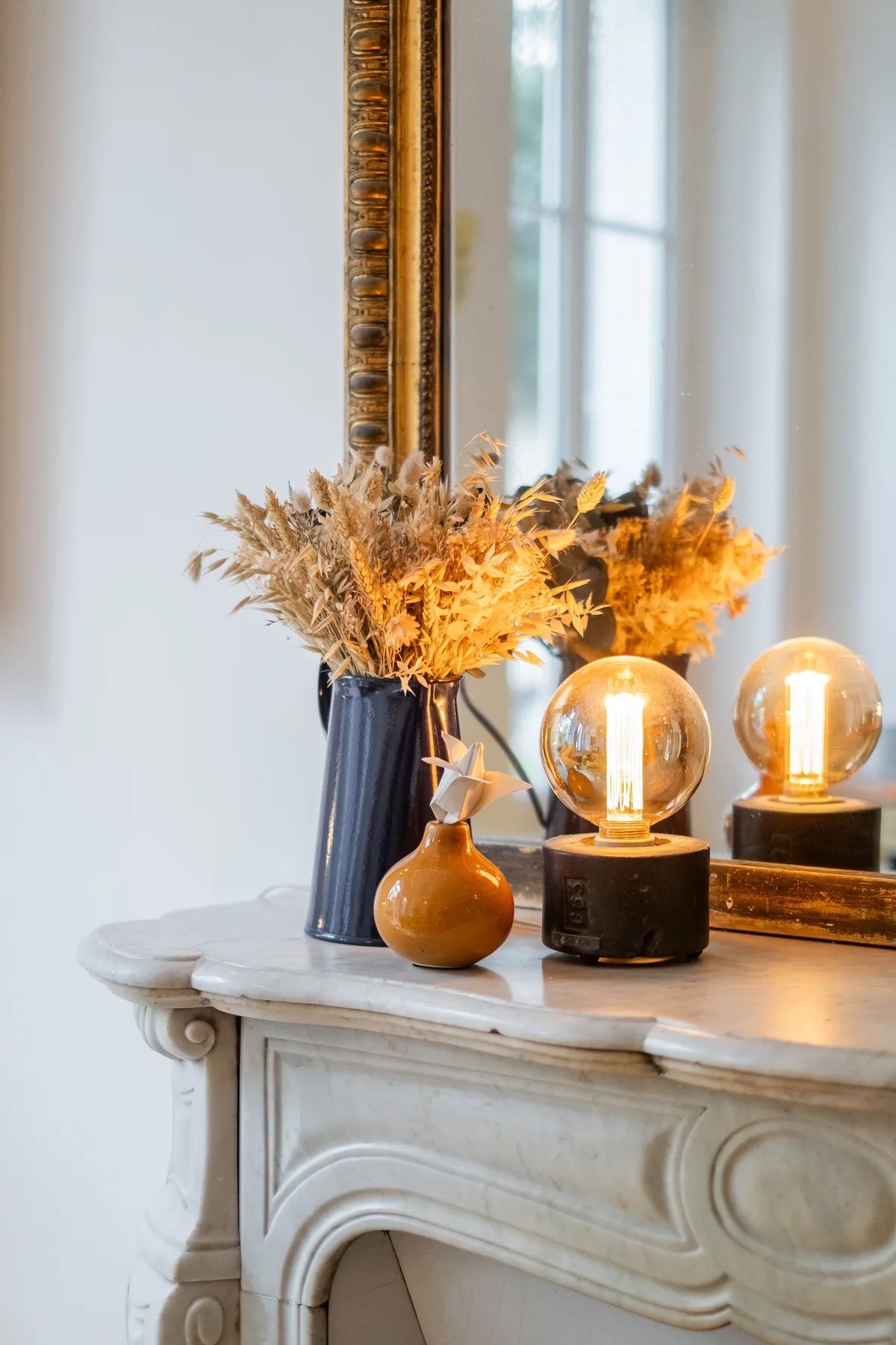 A decorative mantel displays dried flowers in a vase beside glowing table lamps.