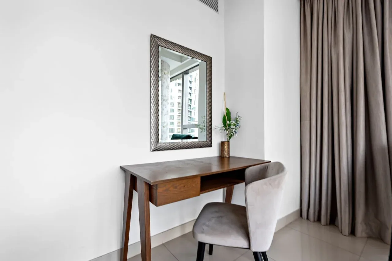 Un escritorio y una silla están junto a una ventana con cortinas, con un espejo sobre el escritorio.