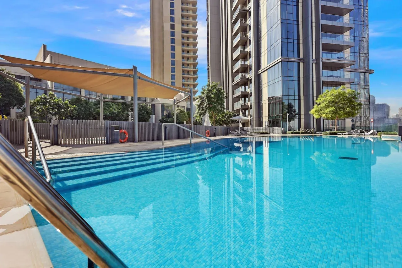 Una piscina exterior junto a edificios modernos de gran altura, con una zona de estar sombreada.