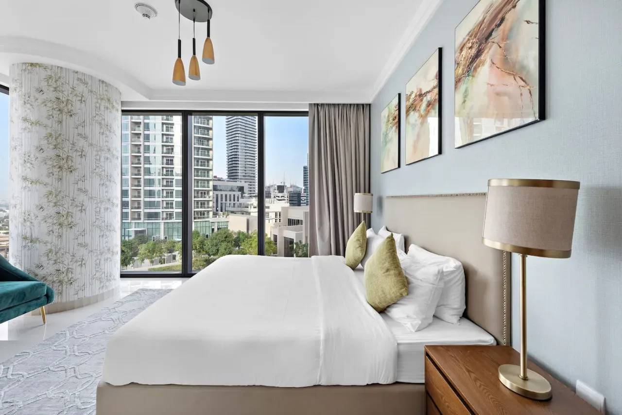 A bright bedroom features a neatly made bed beside large windows overlooking city buildings.