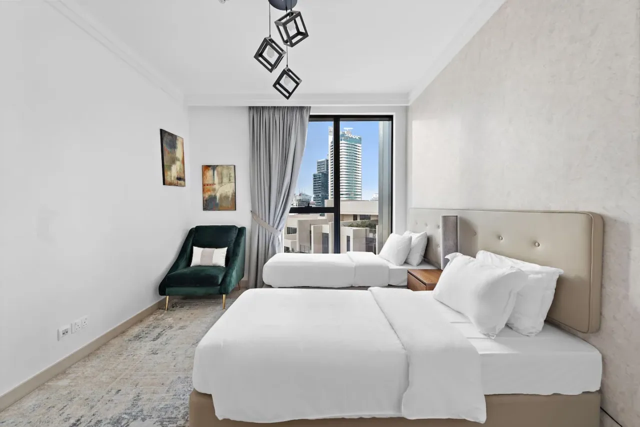 Un dormitorio luminoso con ropa de cama blanca, un sillón verde y una ventana amplia.