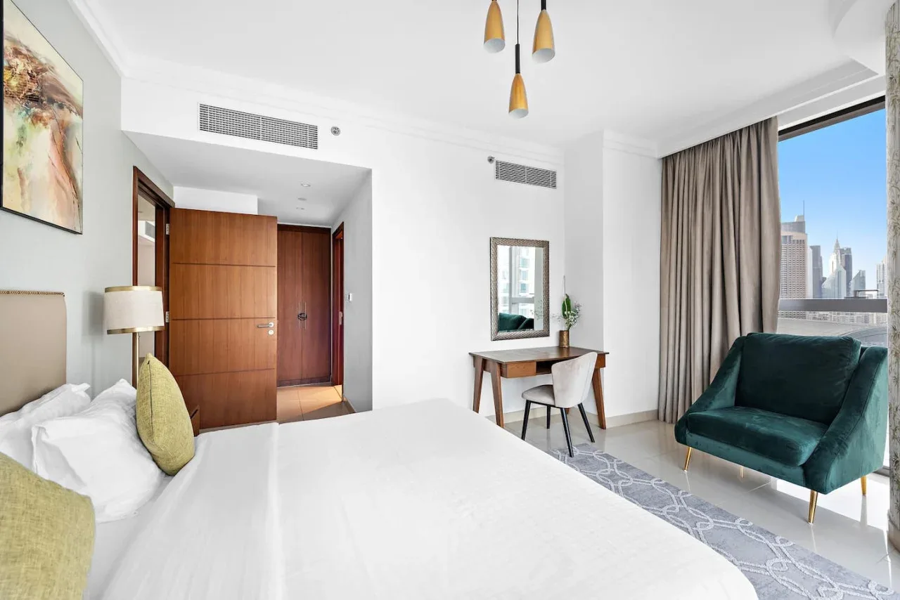 Un dormitorio luminoso con una cama, un escritorio y un sillón verde azulado junto a la ventana.