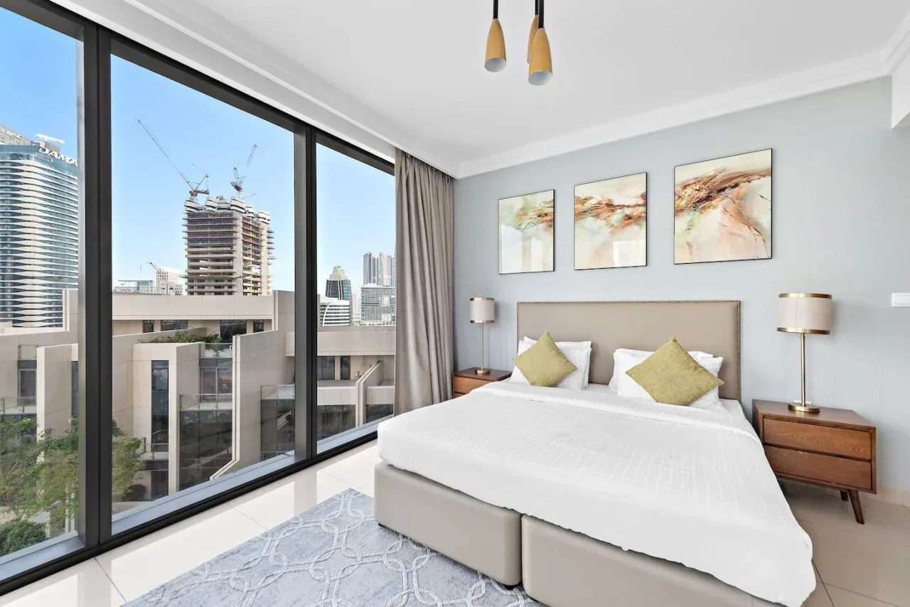 Un dormitorio luminoso tiene una cama grande junto a ventanales con vistas a edificios cercanos.