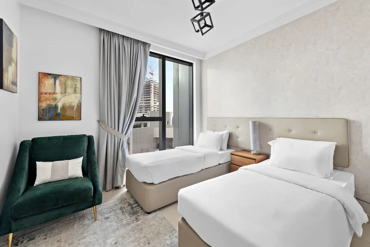 Un dormitorio luminoso con dos camas, un sillón verde y una ventana grande.