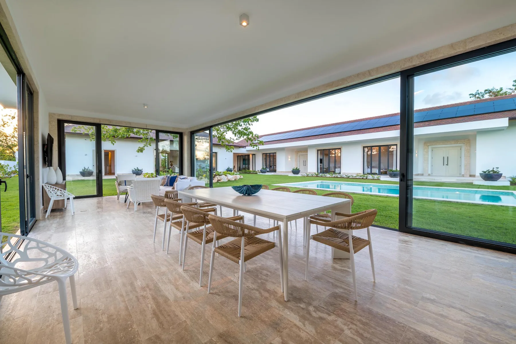 A dining area with a long table and seating overlooks a garden and pool.