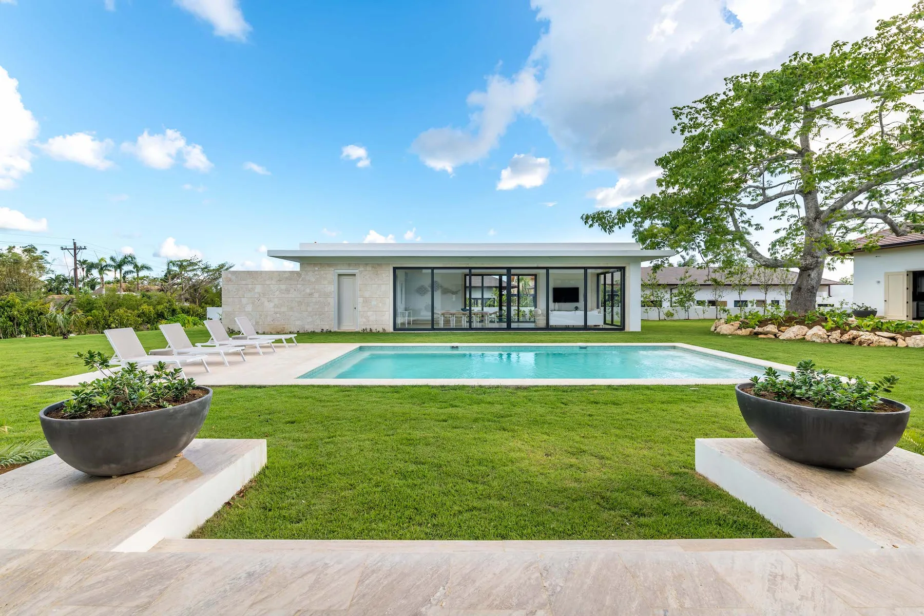 A modern house faces a swimming pool, with lounge seating on the lawn.