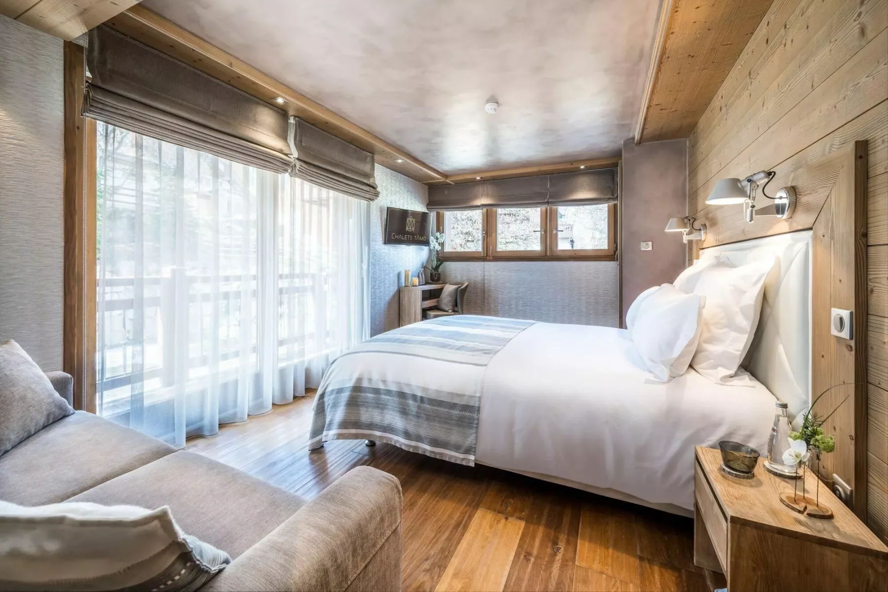A bedroom with a neatly made bed, large windows, and wood-paneled walls.
