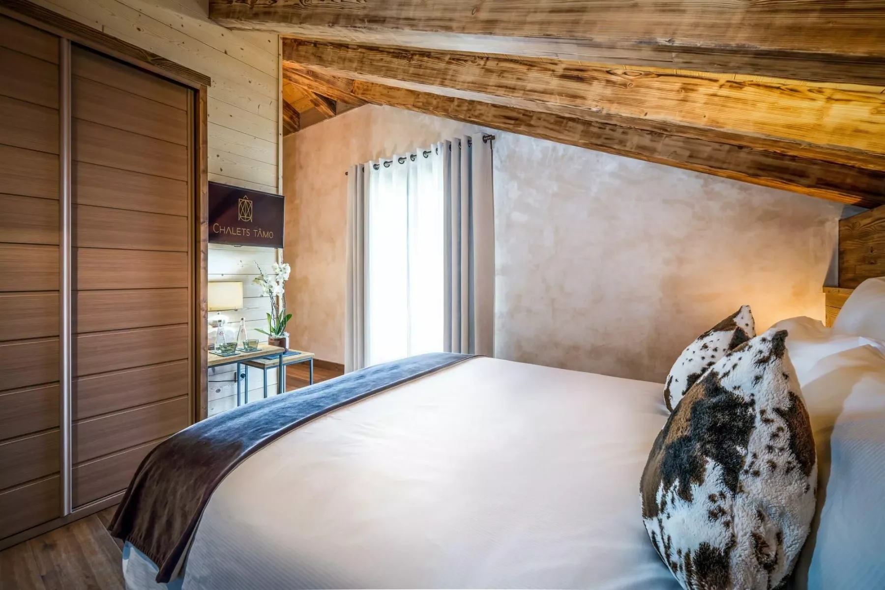 A bedroom with a large bed and a curtained window beneath exposed wooden beams.