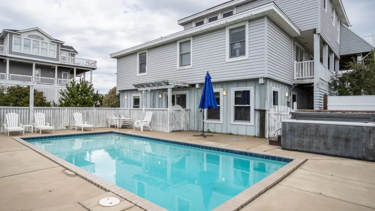 Una piscina está junto a una casa grande de color claro y un patio pavimentado.