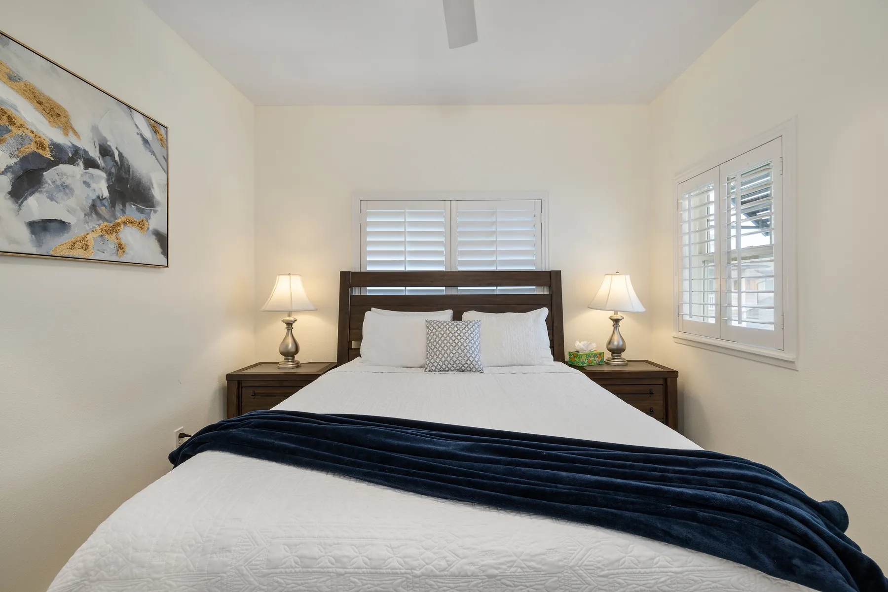 Un dormitorio con una cama tendida y ventanas con persianas blancas.