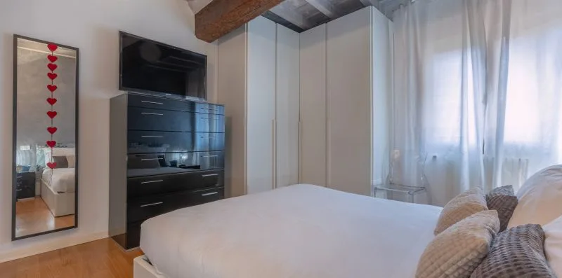 Un dormitorio con una cama, una cómoda oscura y una ventana con cortinas translúcidas.