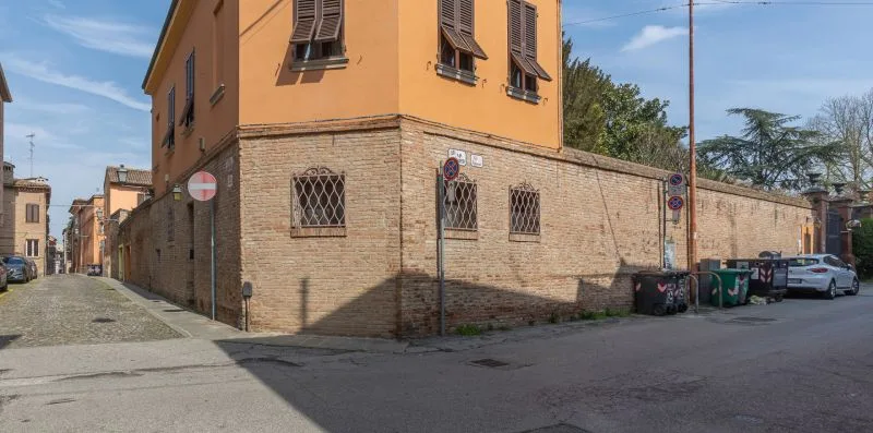 Una esquina junto a un edificio naranja, con coches aparcados y contenedores junto al muro.
