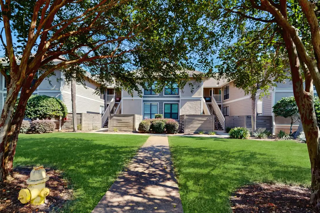 A residential building stands beyond a paved path crossing a green lawn.