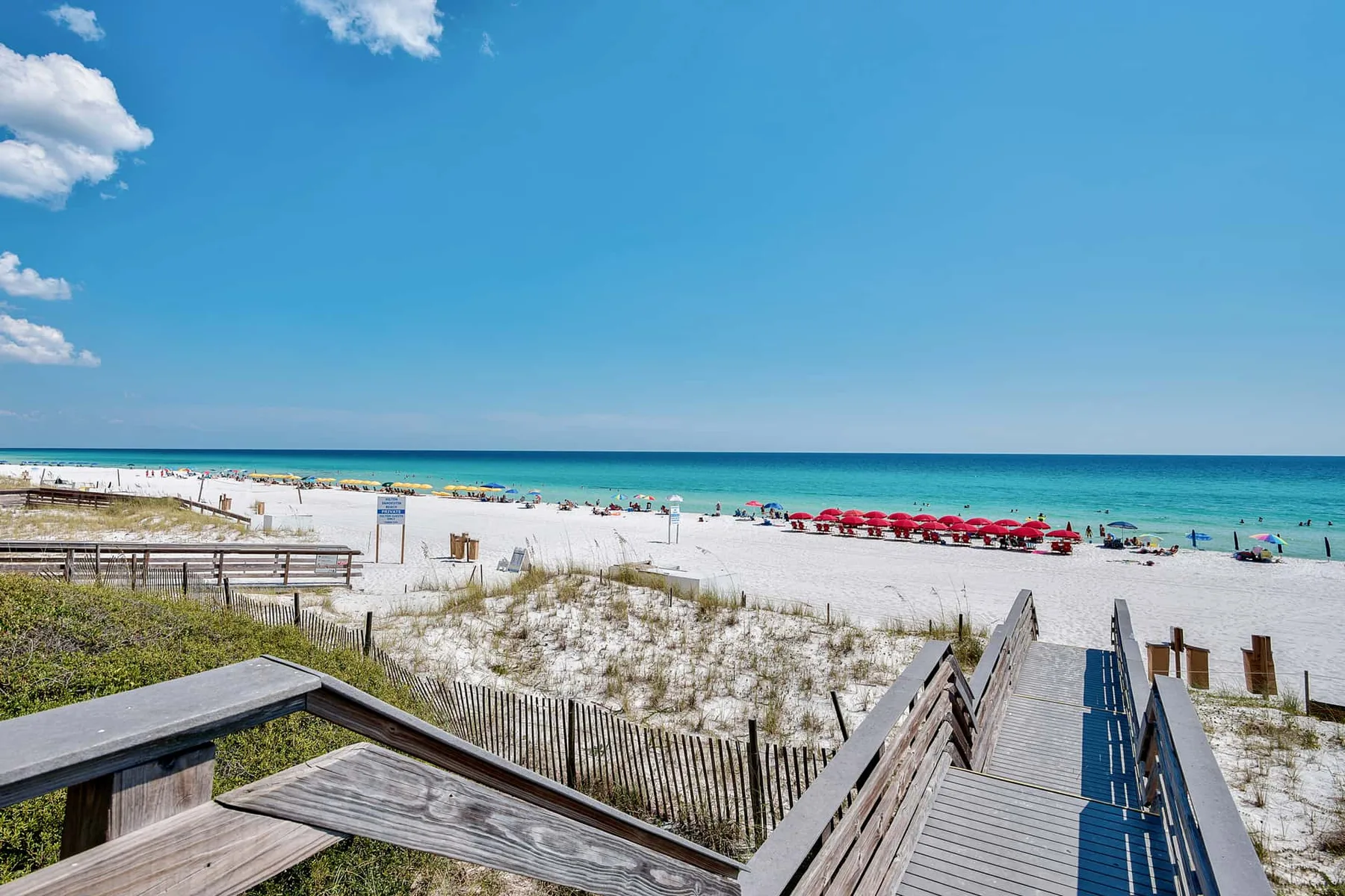 A wooden boardwalk crosses sandy dunes toward a beach with red umbrellas.