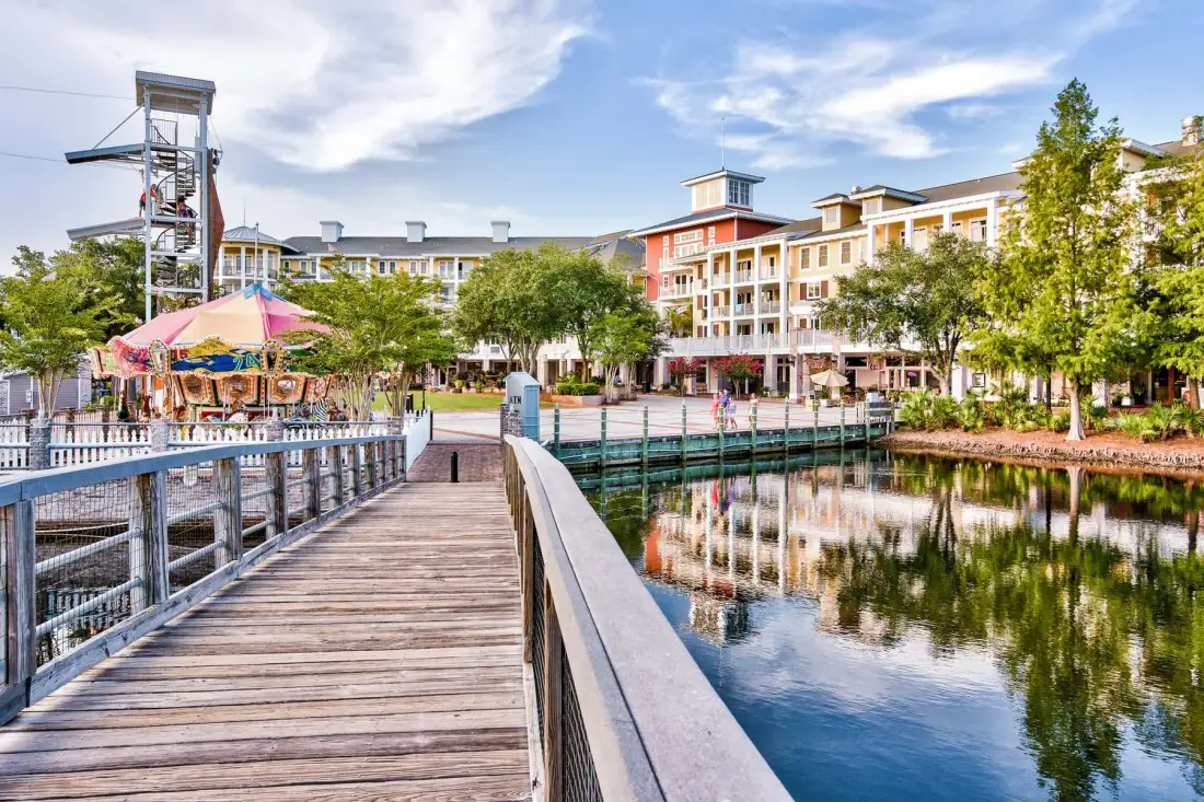 A wooden boardwalk crosses a waterfront beside trees and multistory buildings.