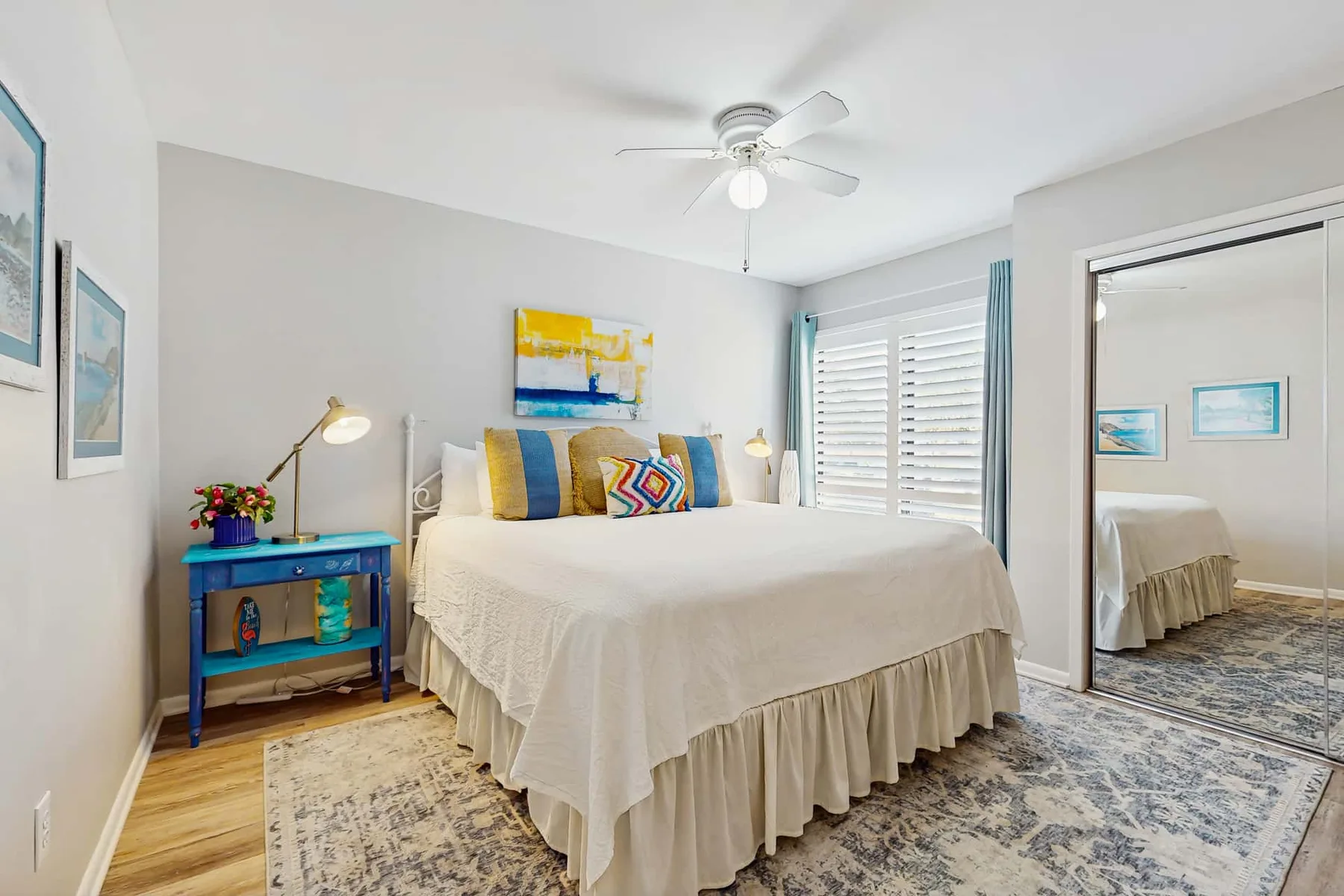 A bright bedroom features a large bed, colorful pillows, and a ceiling fan.