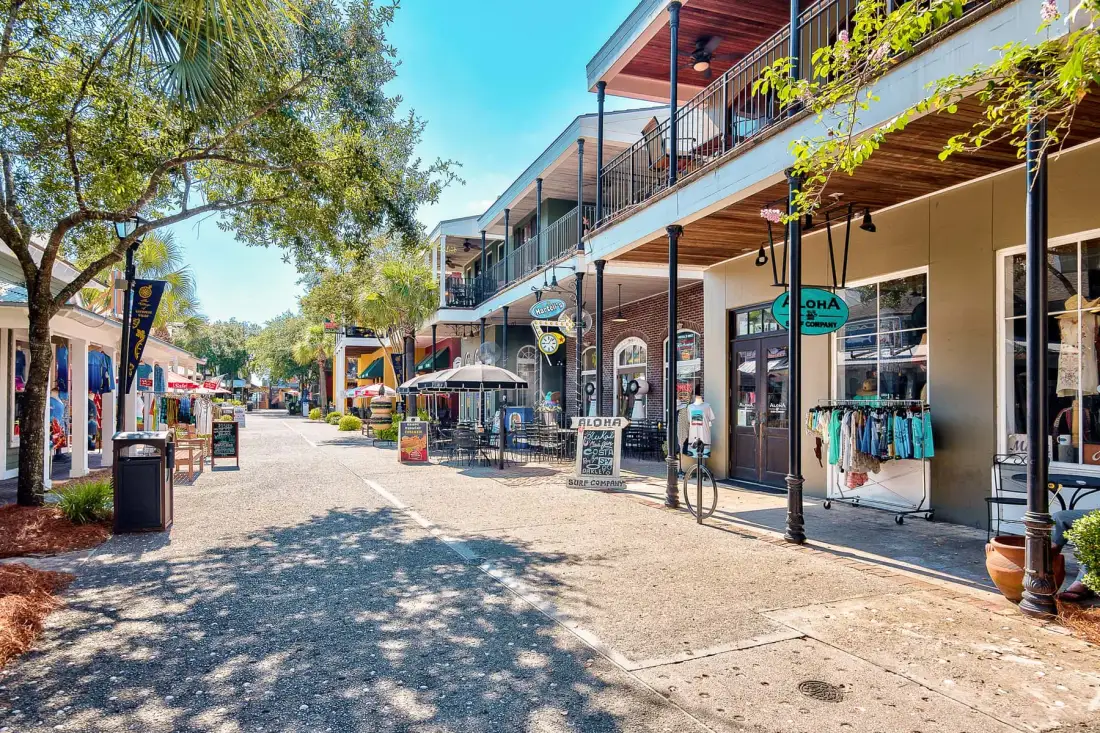 A sunny pedestrian shopping street with storefronts, outdoor seating, and leafy trees.