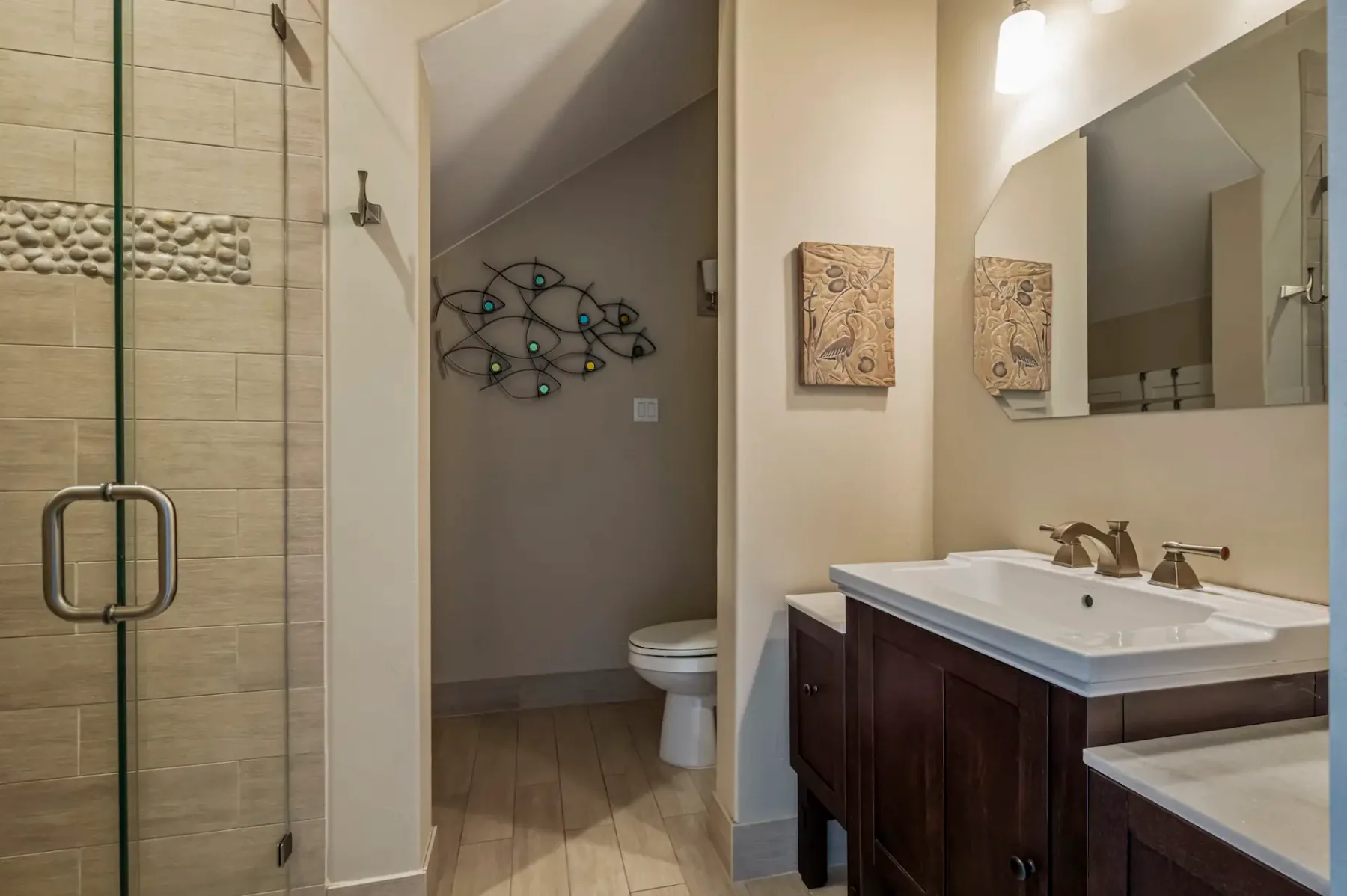 Modern bathroom with glass-enclosed shower, dark wood vanity, rectangular mirror, and decorative wall art above toilet.