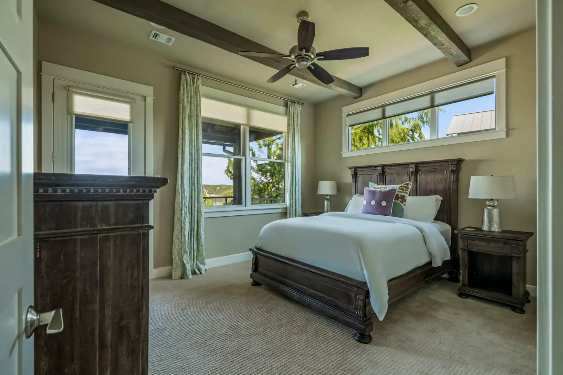 Cozy bedroom with a large bed, wooden furniture, ceiling fan, and windows with curtains showing an outdoor view. Neutral color palette.