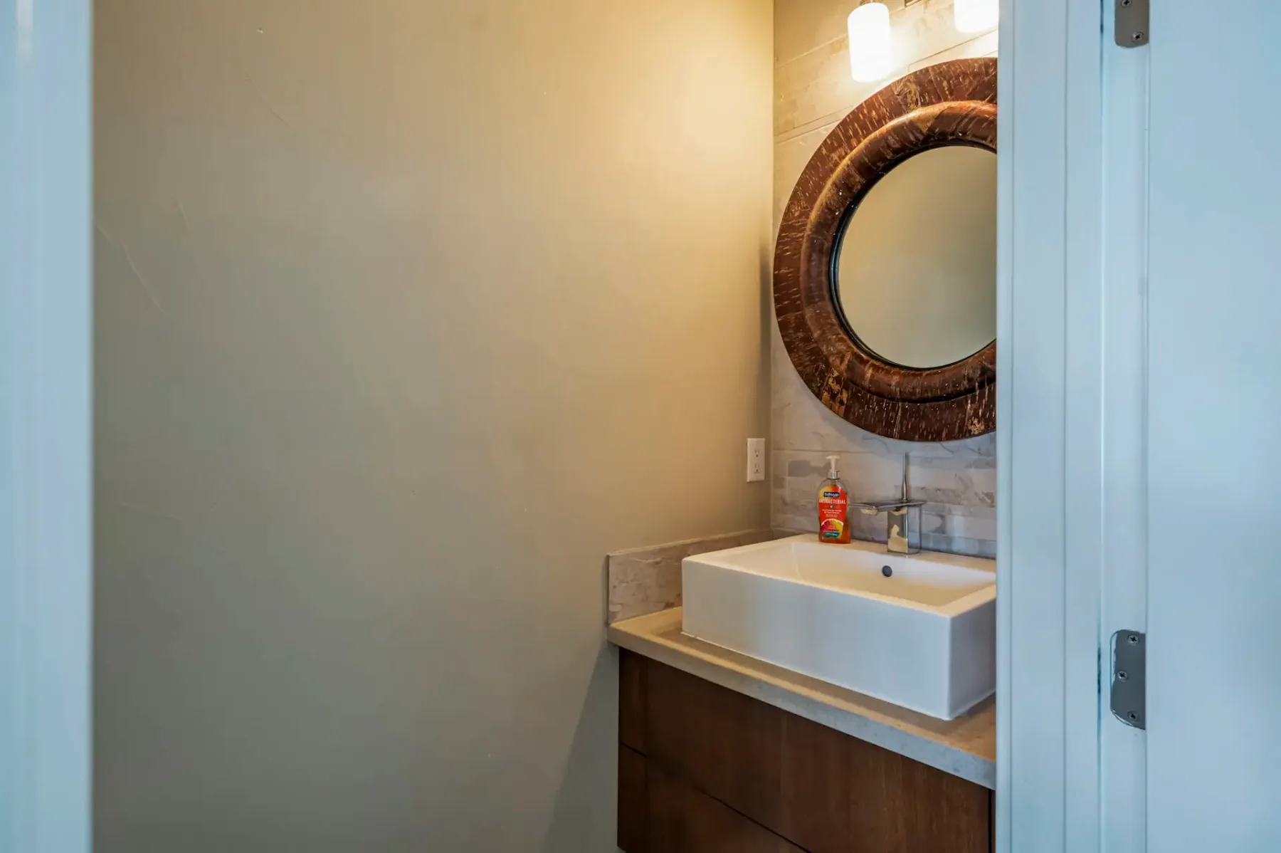 Small bathroom with a round mirror, a vessel sink, and a soap dispenser on a wooden vanity. Soft lighting from a wall fixture above.