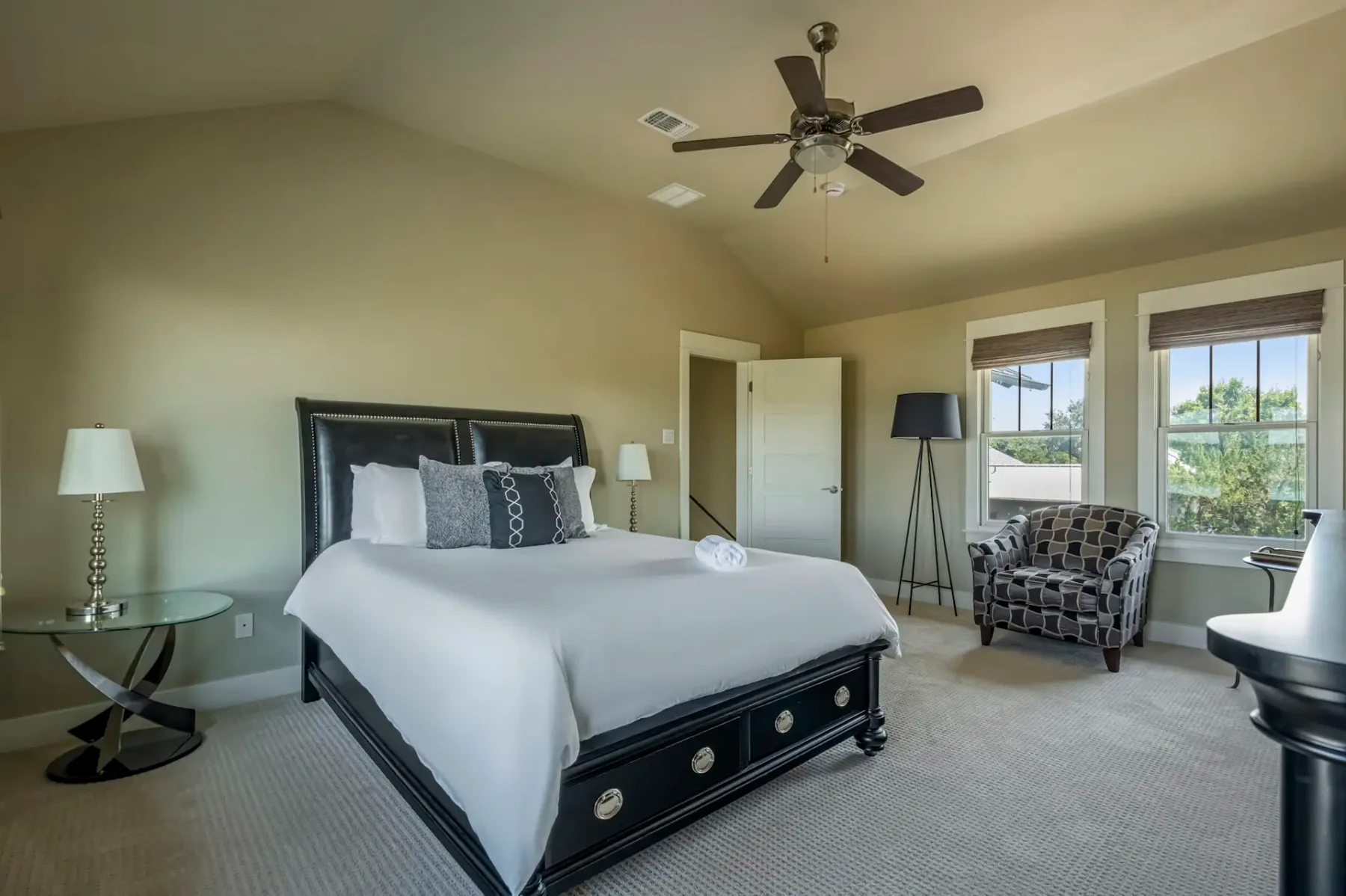 Bedroom with a large bed, black headboard, and white bedding. Ceiling fan, round side tables, patterned armchair, and floor lamp near windows.