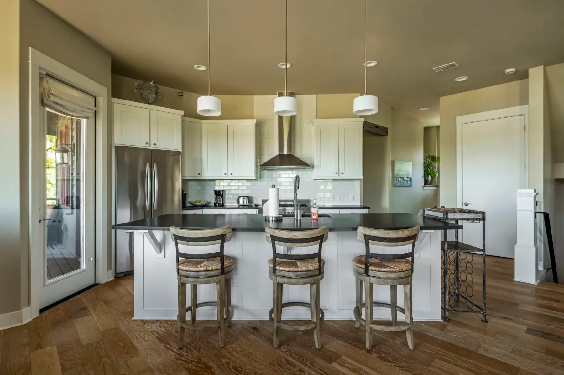 Modern kitchen with a large island, three wooden barstools, stainless steel appliances, and white cabinetry, on hardwood flooring.