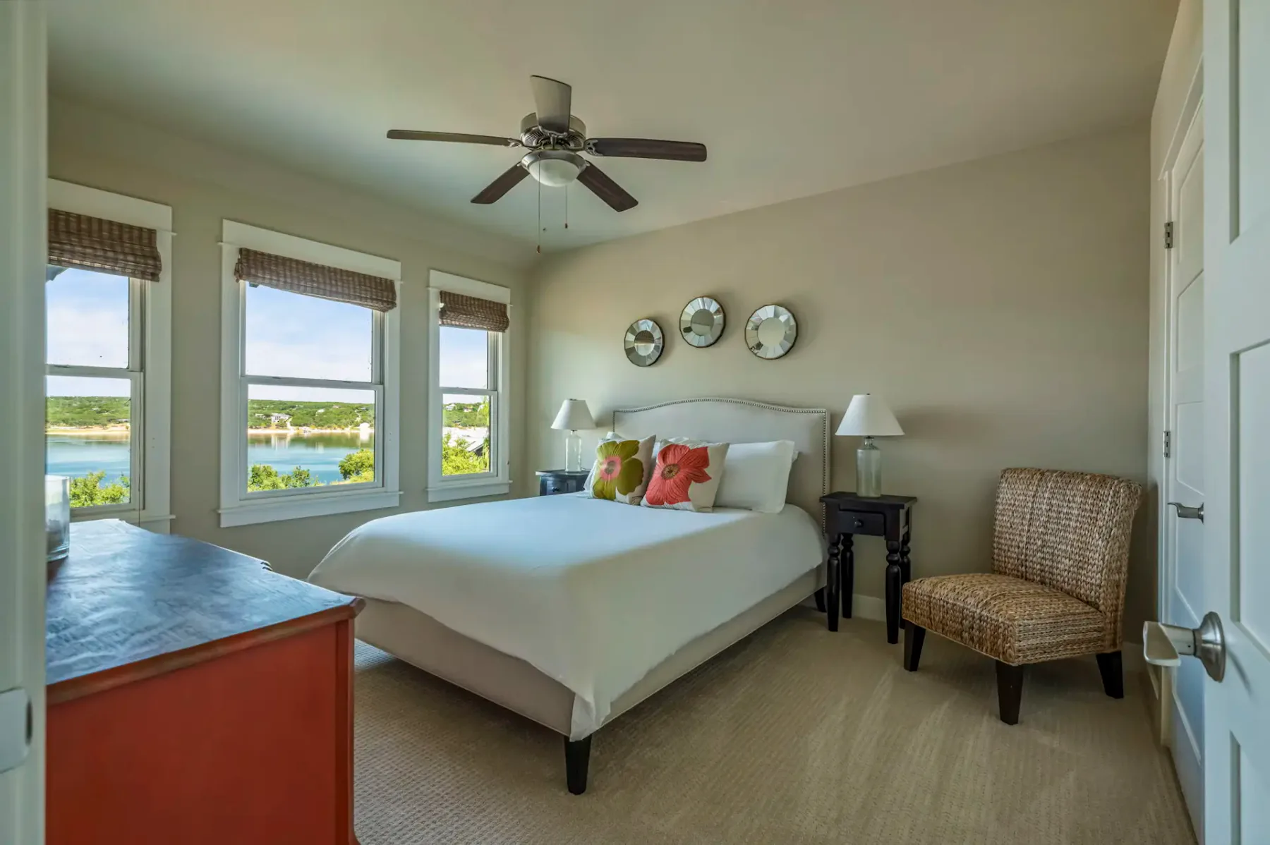 Cozy bedroom with a large bed, colorful pillows, wicker chair, ceiling fan, three wall plates, and windows offering a scenic view of a lake.