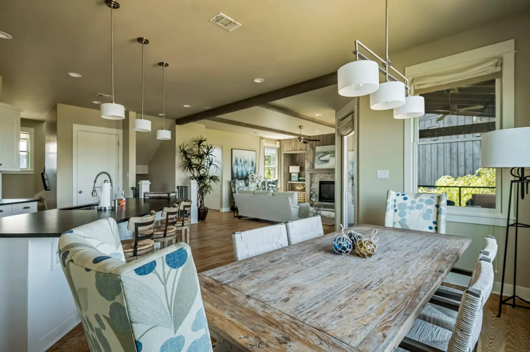 Open-concept dining and living area with rustic wooden table, upholstered chairs, modern lighting, and neutral-toned decor. Large windows offer natural light.