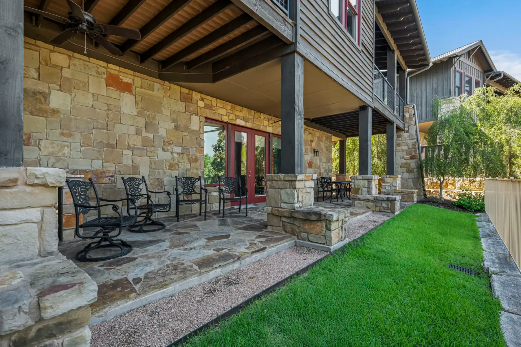 Stone patio with wrought iron chairs, a fan overhead, and a garden view. Two-story house with wood and stone exterior in the background.