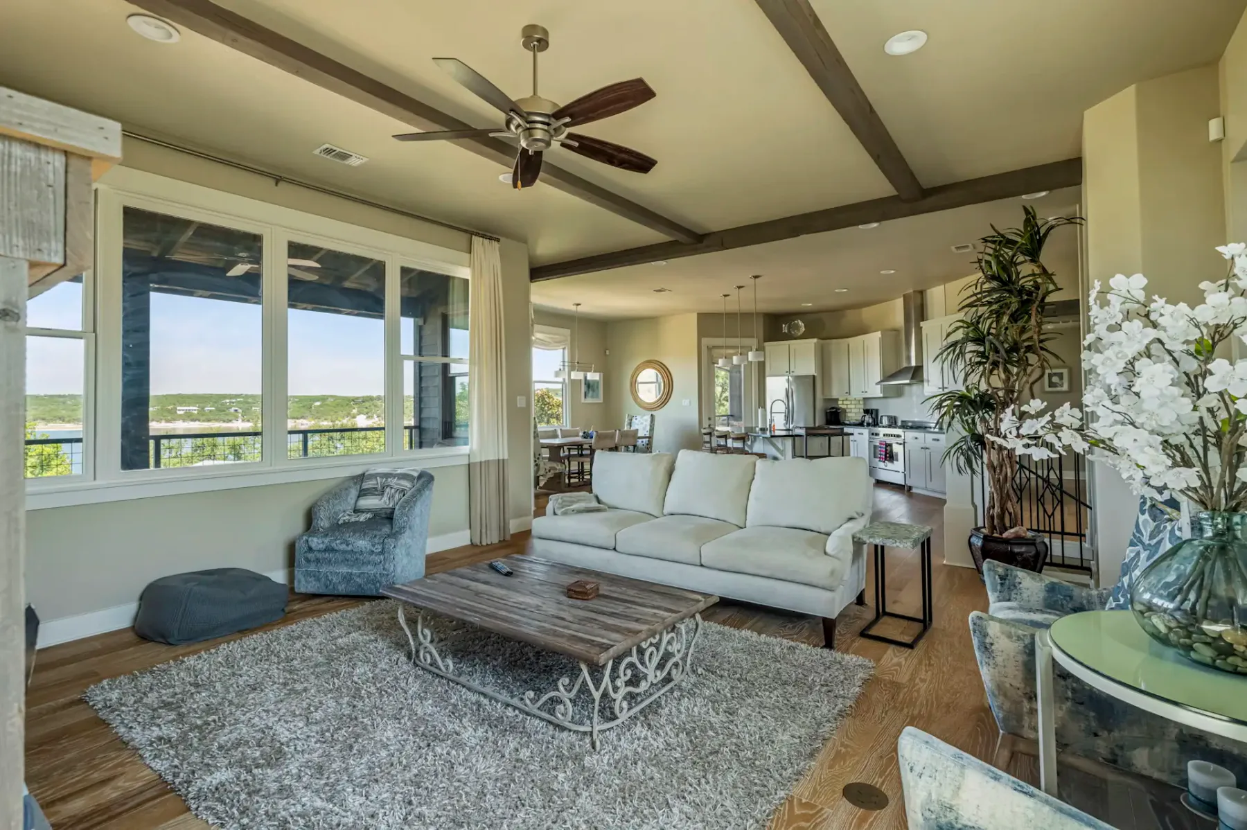 Open-concept living room with beige sofa, wooden coffee table, and kitchen in background. Large window with scenic view, ceiling fan, and potted plants.