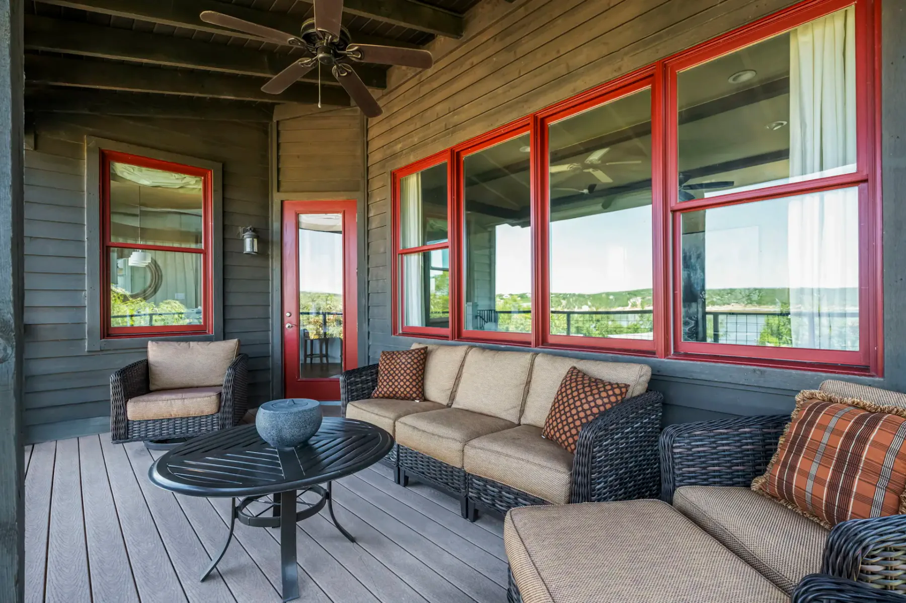 A covered porch with wicker seating and red-trimmed windows overlooking greenery.