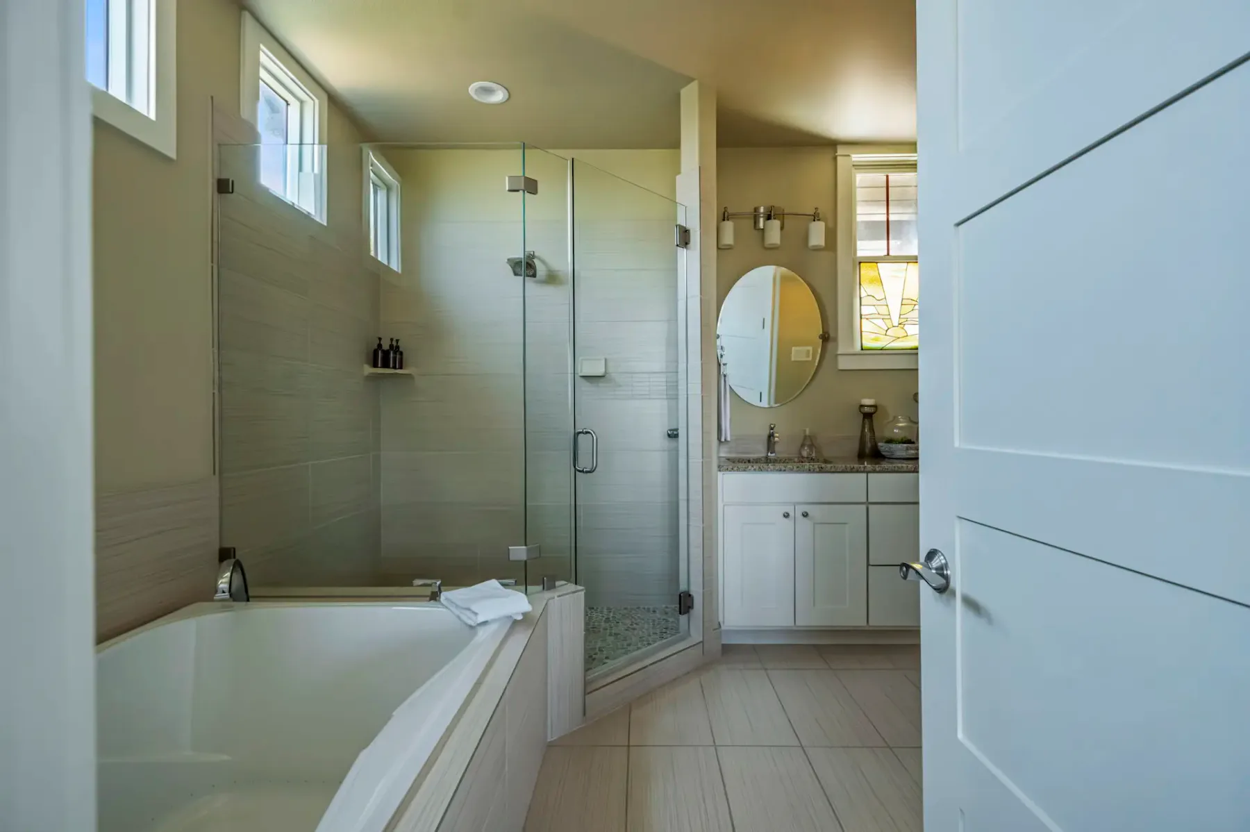 Modern bathroom with a glass shower, bathtub, oval mirror, and white cabinets. Natural light streams through small windows above.