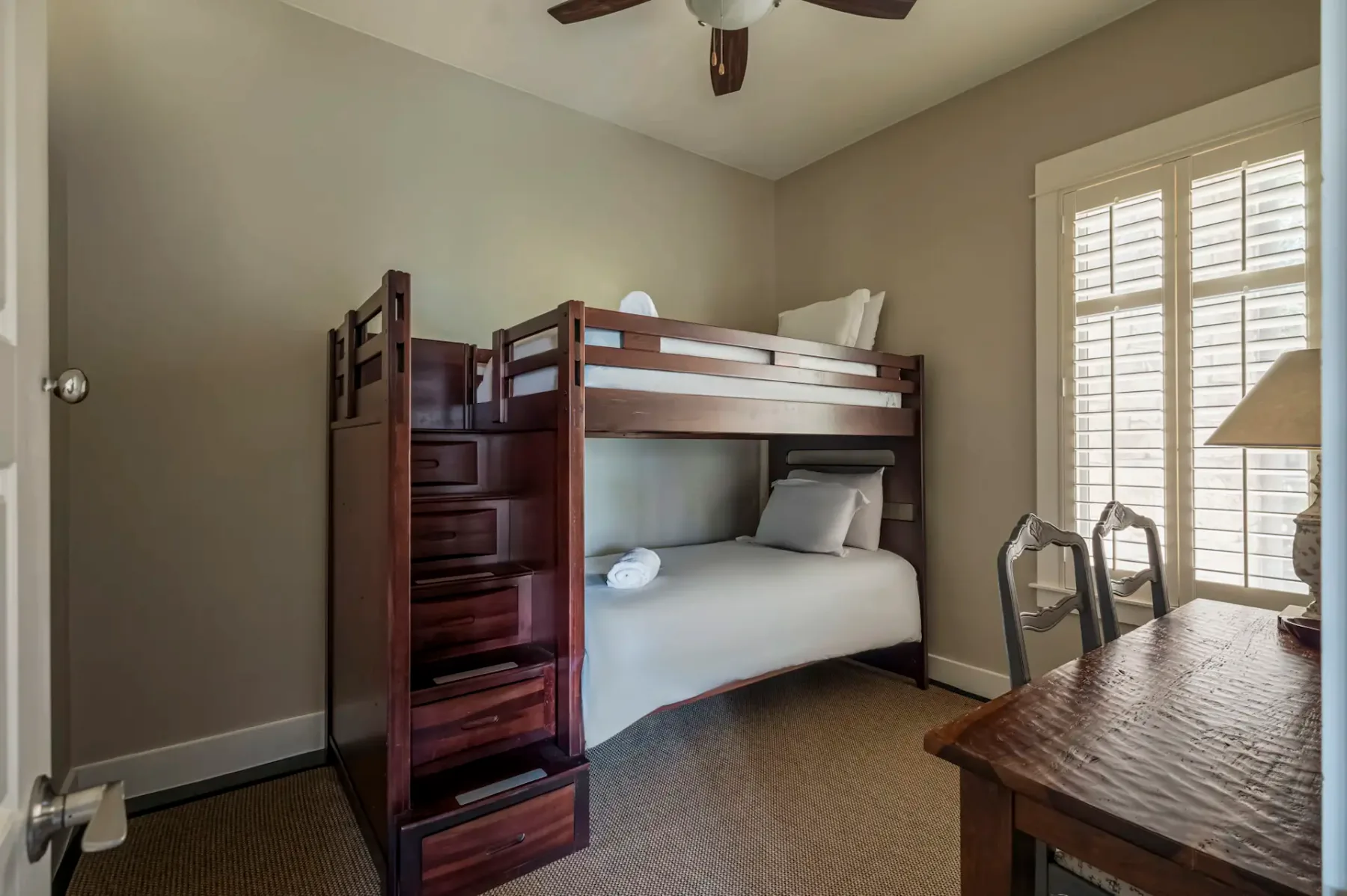 Bunk bed with stairs, white bedding, and pillows in a small room with beige walls, carpeted floor, wooden desk, chairs, and a window with shutters.