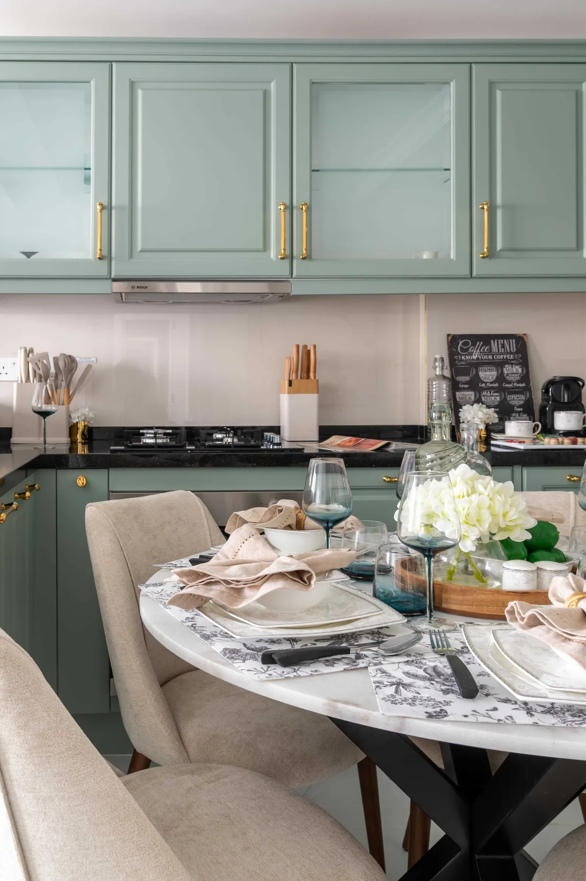 Una mesa puesta con vajilla está junto a una cocina con gabinetes verdes.