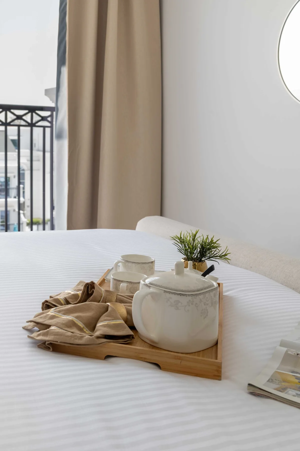 Una cama bien arreglada tiene una bandeja de madera con una tetera, tazas y una planta pequeña.