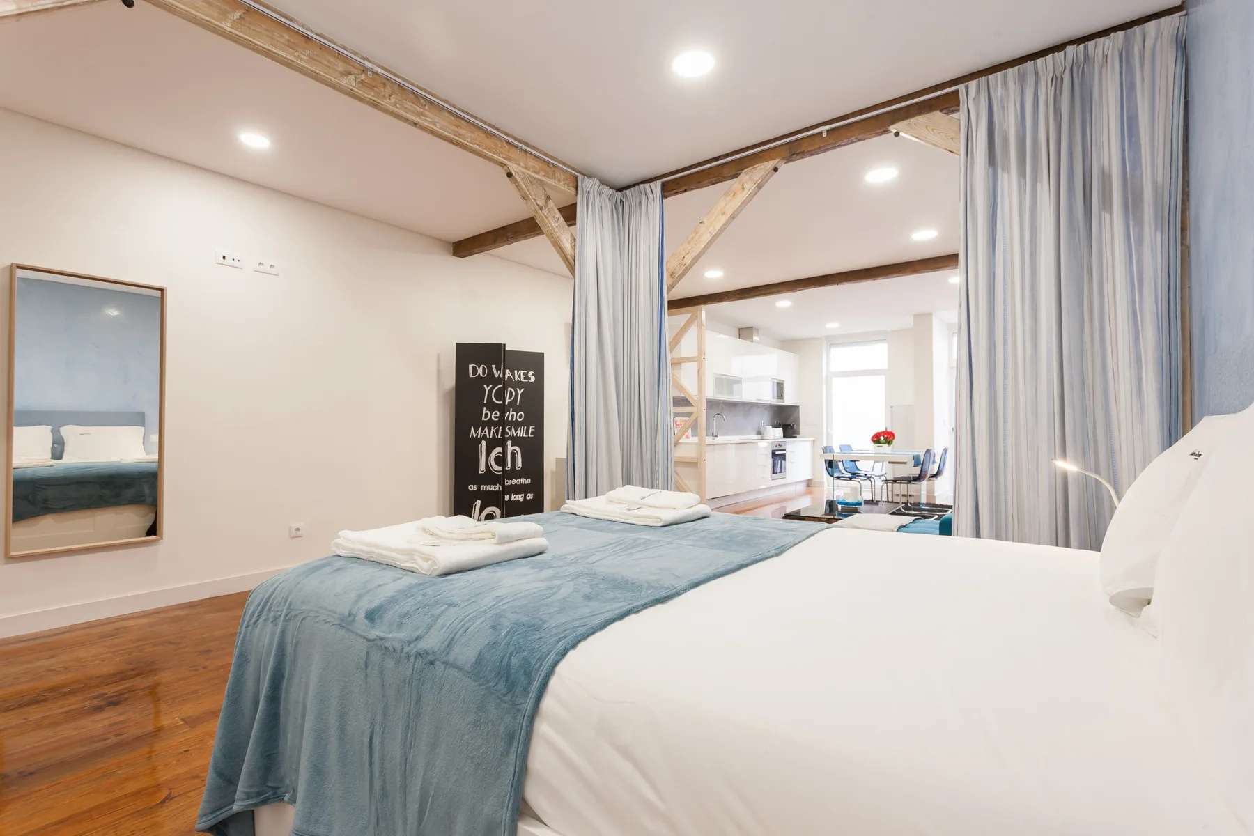 Un dormitorio con una cama y ropa de cama azul, abierto a una luminosa sala de estar.