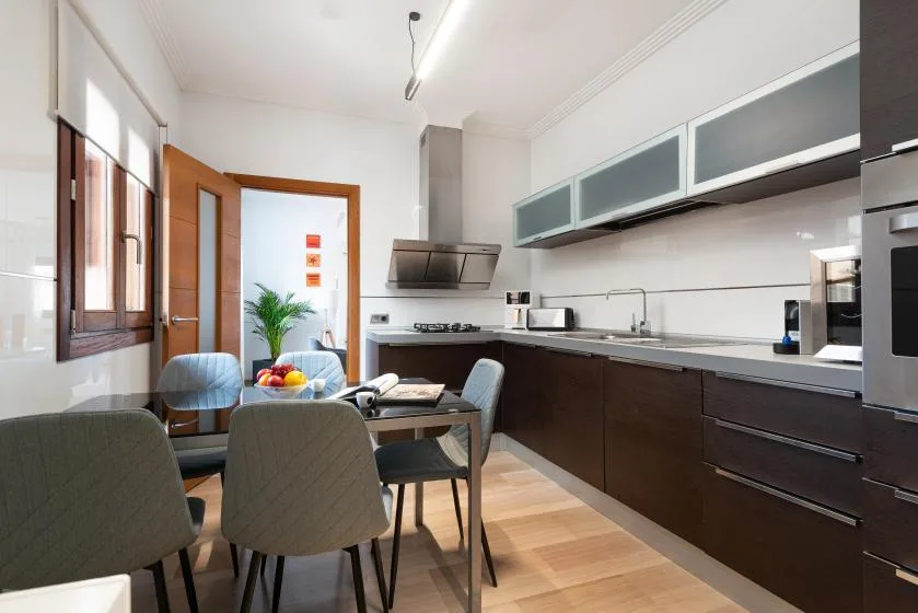 Una cocina y comedor luminosos con gabinetes oscuros, mesa y sillas.