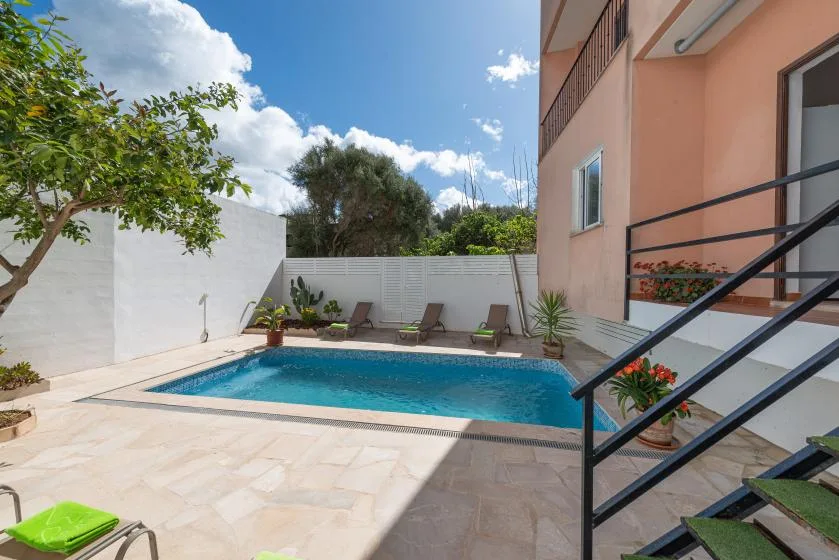 Una piscina exterior soleada está junto a un edificio color durazno y un patio pavimentado.