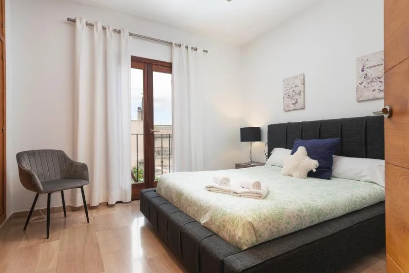Un dormitorio luminoso con una cama grande, cortinas claras y una silla junto a la ventana.